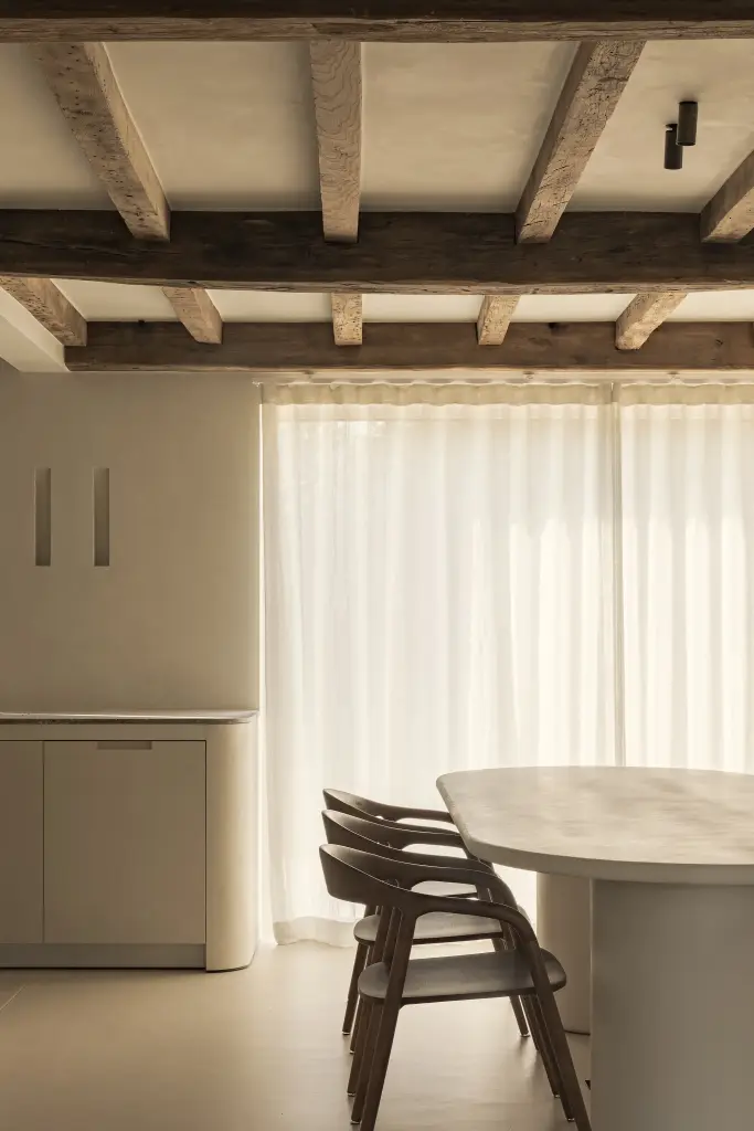 Ovale eettafel met houten stoelen voor linnen gordijnen onder een balkenplafond, Totaalrenovatie in Loppem, interieur Laura Calleeuw